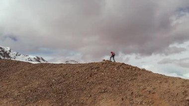 Sırt çantası ve yürüyüş sopalarıyla genç ve zayıf bir adamın havadan görünüşü kayaların ve kayaların sınırındaki tepeye tırmanıyor. Kar örtülü tepe ve bulutların arka planında karla kaplı.