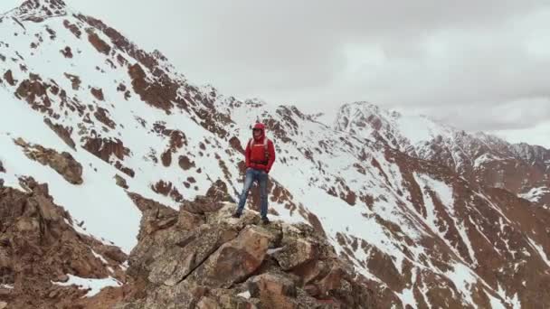 Um viajante do sexo masculino com uma mochila fica no topo de um penhasco contra o pano de fundo de picos montanhosos cobertos de neve e nuvens baixas. Vista aérea. Voando a câmera em torno de um homem em um penhasco de tempestades — Vídeo de Stock