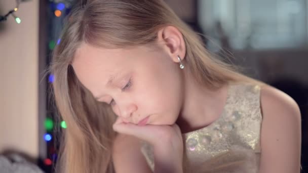 Uma menina em um vestido festivo contra o pano de fundo de luzes festivas e uma árvore de Natal, infelizmente, olha em algum lugar abaixo do borrão e movimento para se concentrar. O conceito de umas férias estragadas — Vídeo de Stock