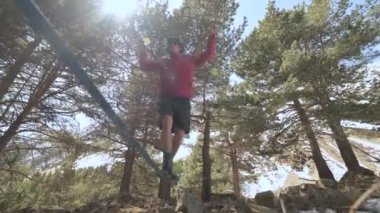 Şort ceketli ve güneş gözlüklü genç bir adam öğleden sonra kozalaklı ormanda gerilmiş bir ip üzerinde dengede duruyor. Açık hava sporları ve eğlence konsepti