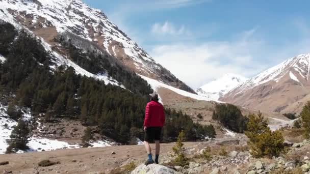 Una vista aérea de un joven con gorra y gafas de sol y pantalones cortos se encuentra en las montañas y mira los picos nevados. El concepto de turismo de montaña y viajes en gras — Vídeos de Stock