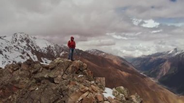 Sırt çantalı bir erkek yolcu bir uçurumun tepesinde karlı dağ zirveleri ve alçak bulutların zemininde duruyor. Hava görüntüsü. Storon uçurumundaki bir adamın etrafında kamerayı uçurmak