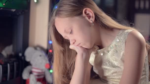 Ein kleines Mädchen in einem festlichen Kleid vor dem Hintergrund festlicher Lichter und eines Weihnachtsbaums blickt traurig durch die Unschärfe und Bewegung, um sich zu fokussieren. Das Konzept eines verwöhnten Urlaubs — Stockvideo