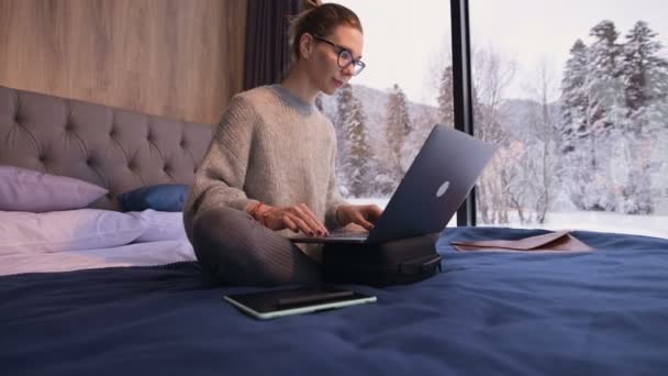 Девушка с собранными волосами в очках сидит на кровати с ноутбуком в руках против панорамных окон, за которыми зимний лес в снегу. — стоковое видео