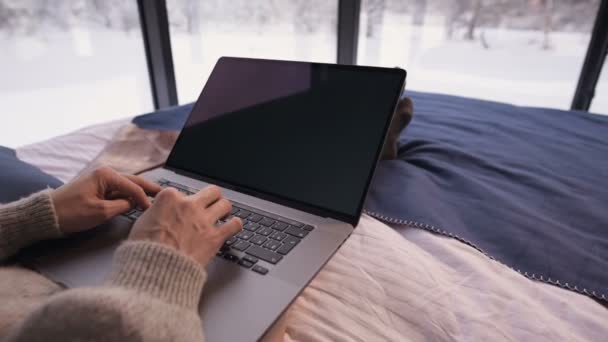 Close-up das mãos da menina imprimir algo no laptop se senta em uma cama com um laptop em suas mãos contra as janelas panorâmicas atrás das quais a floresta de inverno na neve. — Vídeo de Stock