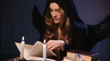 Orta boy, beyaz, siyah başlıklı, altın sarısı, uzun saçlı bir kız bir masada oturuyor ve kitaptan yapraklar döküyor. Kanatlı kostüm. Mumlar yanıyor. Cadılar Bayramı konsepti