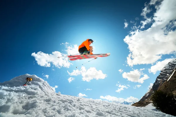 Um jovem homem elegante em óculos de sol e um boné executa um truque em saltar com um chute de neve contra o céu azul e o sol em um dia ensolarado. O conceito de esqui no parque no inverno ou na primavera — Fotografia de Stock