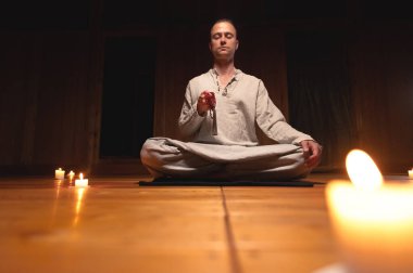 Antrenman ve meditasyon için giyinmiş bir adam lotus pozisyonunda oturur ve loş ışıklı ahşap bir odada dikkat çekmek için kırmızı bir tespih tutar.