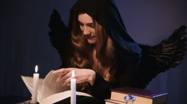 Orta boy, beyaz, siyah başlıklı, altın sarısı, uzun saçlı bir kız bir masada oturuyor ve kitaptan yapraklar döküyor. Kanatlı kostüm. Mumlar yanıyor. Cadılar Bayramı konsepti