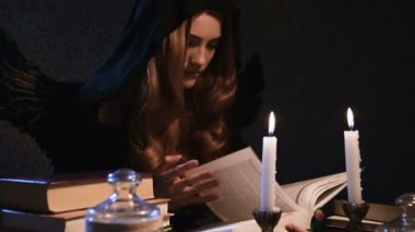 Orta boy, beyaz, siyah başlıklı, altın sarısı, uzun saçlı bir kız bir masada oturuyor ve kitaptan yapraklar döküyor. Kanatlı kostüm. Mumlar yanıyor. Cadılar Bayramı konsepti