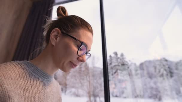Крупным планом девушка с собранными волосами в очках сидит на кровати с ноутбуком в руках против панорамных окон, за которыми зимний лес в снегу. — стоковое видео