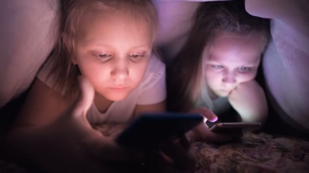 Две остроумные девочки играют ночью на смартфоне под одеялом. Чайлдс уходит от реальности через мобильные устройства. Ребенок пользуется секретным телефоном — стоковое видео