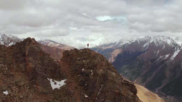 Een mannelijke reiziger met een rugzak staat bovenop een klif tegen de achtergrond van besneeuwde bergtoppen en lage wolken. Luchtfoto 's. De camera rond een man in een stormklif vliegen — Stockvideo