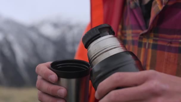 Um homem de close-up derrama chá quente de uma garrafa térmica em uma caneca contra as montanhas cobertas de neve do Cáucaso. O conceito de calor e conforto em viagens . — Vídeo de Stock