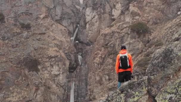 Luftaufnahme eines jungen männlichen Fotografen mit einer Kamera in der Hand steht auf einem hohen Felsen in einer Schlucht und fotografiert einen alpinen Wasserfall bei Schneesturm und Schneefall im Frühling. — Stockvideo