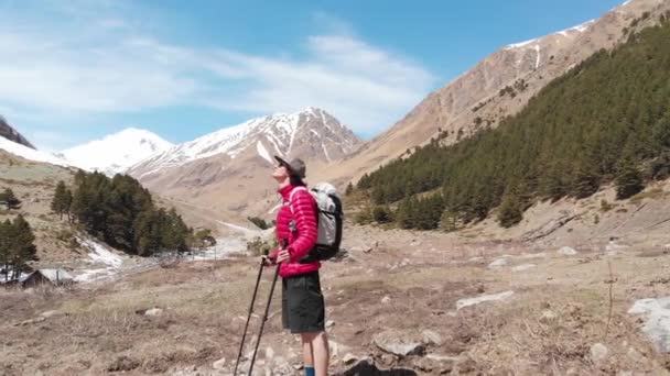 산에는 모자를 쓰고 선글라스와 반바지를 입은 젊은 남자가 눈덮인 산봉우리를 바라보고 있다. 산악 관광과 그라스 여행의 개념 — 비디오