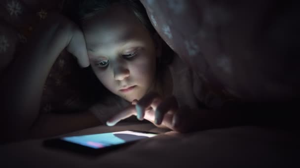 熱心な少女は毛布の下で夜にスマートフォンで再生されます。子供たちは現実からモバイルデバイスに移行します。子供は秘密の電話を使う — ストック動画