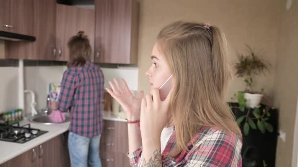 Missnöjd flicka talar i telefon i köket i bakgrunden av sin pojkvän eller make. — Stockvideo