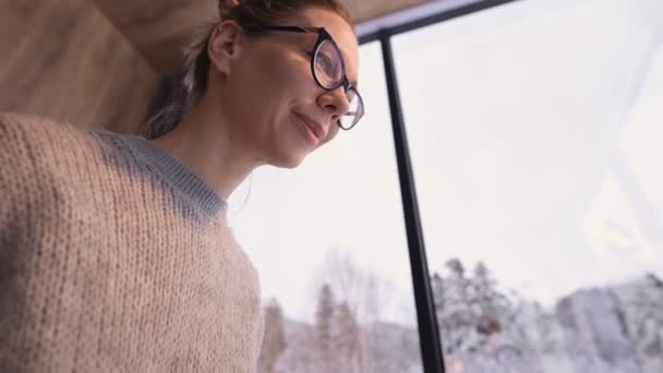 Zblízka dívka s nasbíranými vlasy v brýlích sedí na posteli s notebookem v rukou proti panoramatickým oknům, za kterými zimní les ve sněhu. — Stock video