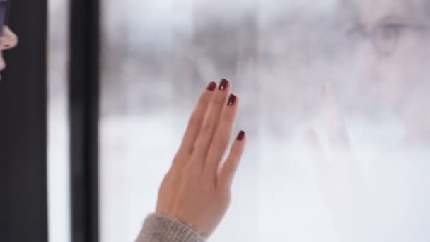 Uma jovem mulher com óculos chega à grande janela panorâmica da casa que fica na floresta de inverno toca a mão de vidro e olha atentamente para a distância. — Vídeo de Stock