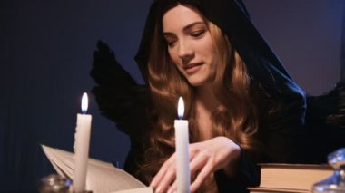 Orta boy, beyaz, siyah başlıklı, altın sarısı, uzun saçlı bir kız bir masada oturuyor ve kitaptan yapraklar döküyor. Kanatlı kostüm. Mumlar yanıyor. Cadılar Bayramı konsepti