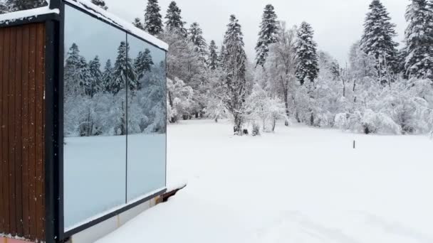 Vista aérea de una innovadora casa de alta tecnología con paredes transparentes espejadas en invierno cerca de un bosque de coníferas en las montañas del Cáucaso o los Alpes. — Vídeos de Stock