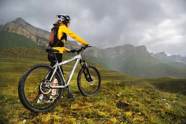 Una chica en un casco y con una mochila con una bicicleta se levanta contra el telón de fondo de montañas épicas y rocas y cielo nublado por la noche. Ciclista mira a las montañas antes de descender mtb —  Fotos de Stock