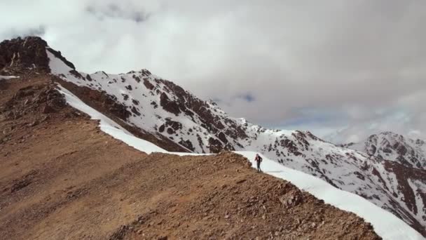 El escalador sube cuesta arriba con una mochila y bastones de trekking en la cresta en el cruce de piedra y nieve. Vista aérea en lo alto de las montañas del Cáucaso norte — Vídeos de Stock