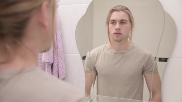 Un joven de pelo largo se acerca al espejo del baño y se mira a sí mismo por la mañana. deja el marco mostrando que está siguiendo la cámara — Vídeos de Stock