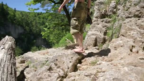 Een man met een kale romp in een strohoed loopt blootsvoets over de rotsen naar de klif om te gaan zitten en te kijken naar de kloof van de rots en het bos. Het concept van alleen reizen — Stockvideo