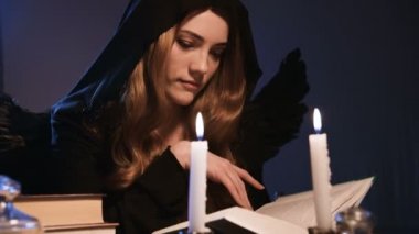 Orta boy, beyaz, siyah başlıklı, altın sarısı, uzun saçlı bir kız bir masada oturuyor ve kitaptan yapraklar döküyor. Kanatlı kostüm. Mumlar yanıyor. Cadılar Bayramı konsepti