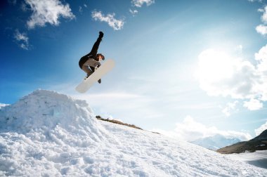 Süslü genç kız snowboardcu, ilkbaharda mavi gökyüzü bulutlarına ve dağlara karşı bir kar topundan atlama numarası yapıyor..