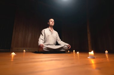 Antrenman ve meditasyon için giyinmiş bir adam lotus pozisyonunda oturur ve loş ışıklı ahşap bir odada dikkat çekmek için kırmızı bir tespih tutar.