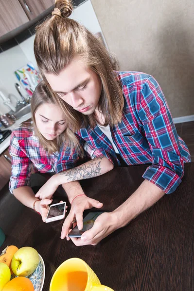 A young smile couple guy and a girl with long hair sit at the dinner table in the kitchen and use phones communicating in chat rooms and social networks. — Stock Photo, Image