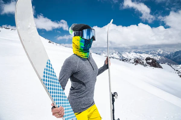 Retrato de uma menina esbelta em um lustre e uma balaclava em uma máscara de esqui e um chapéu com um rosto fechado ao lado de esquis contra o pano de fundo de montanhas épicas cobertas de neve. — Fotografia de Stock