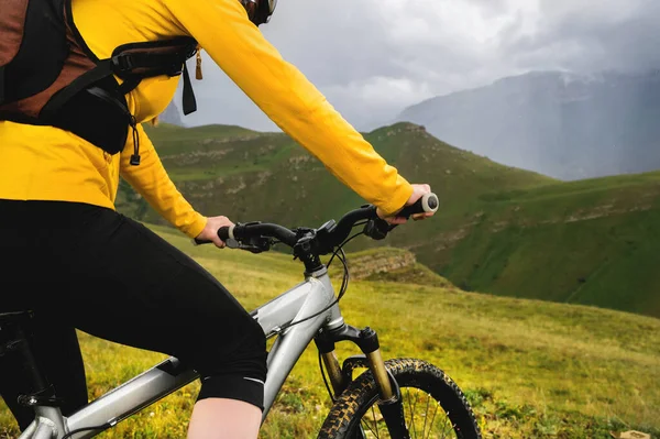 Primer plano joven esbelta chica con una mochila y un casco de bicicleta se sienta en una bicicleta en lo alto de las montañas en un día nublado y mira a las montañas y las nubes . —  Fotos de Stock