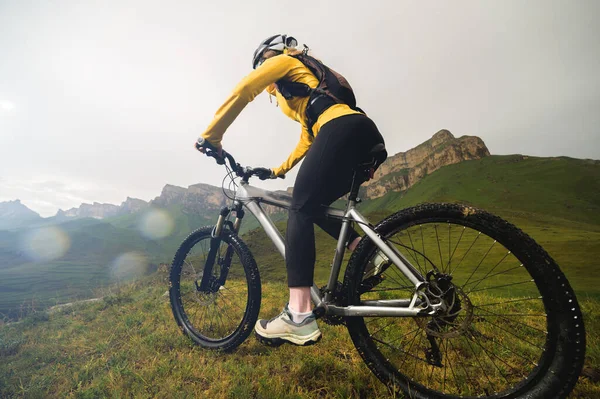 Atractiva chica pelirroja en un casco y con una mochila se sienta en una bicicleta de montaña en las montañas contra el telón de fondo de rocas y cielo nublado —  Fotos de Stock