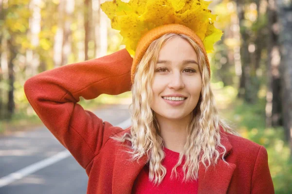 Menina loira atraente com cabelo encaracolado em um boné laranja e casaco vermelho mantém um buquê de folhas amarelas e sorrisos contra a estrada na floresta — Fotografia de Stock