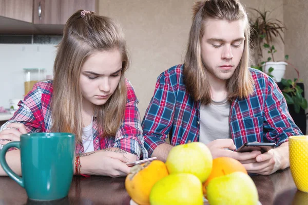 A young smile couple guy and a girl with long hair sit at the dinner table in the kitchen and use phones communicating in chat rooms and social networks. — Stock Photo, Image