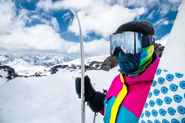 Retrato de uma menina esquiador em uma jaqueta brilhante multi-colorido em uma máscara de esqui com o rosto fechado em um dia ensolarado contra o pano de fundo de montanhas brancas cobertas de neve e nuvens — Fotografia de Stock