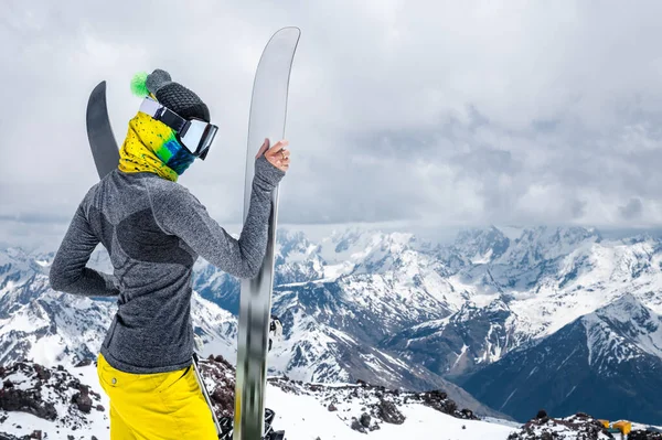 Portret smukłej dziewczyny w opasce i kominiarki w masce narciarskiej oraz kapelusz z zamkniętą twarzą obok nart na tle ośnieżonych epickich gór. — Zdjęcie stockowe