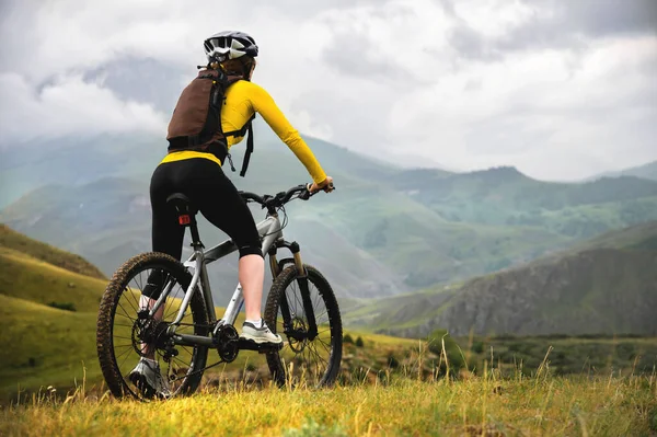 Una joven esbelta con una mochila y un casco de bicicleta se sienta en una bicicleta en lo alto de las montañas en un día nublado y mira las montañas y las nubes . —  Fotos de Stock