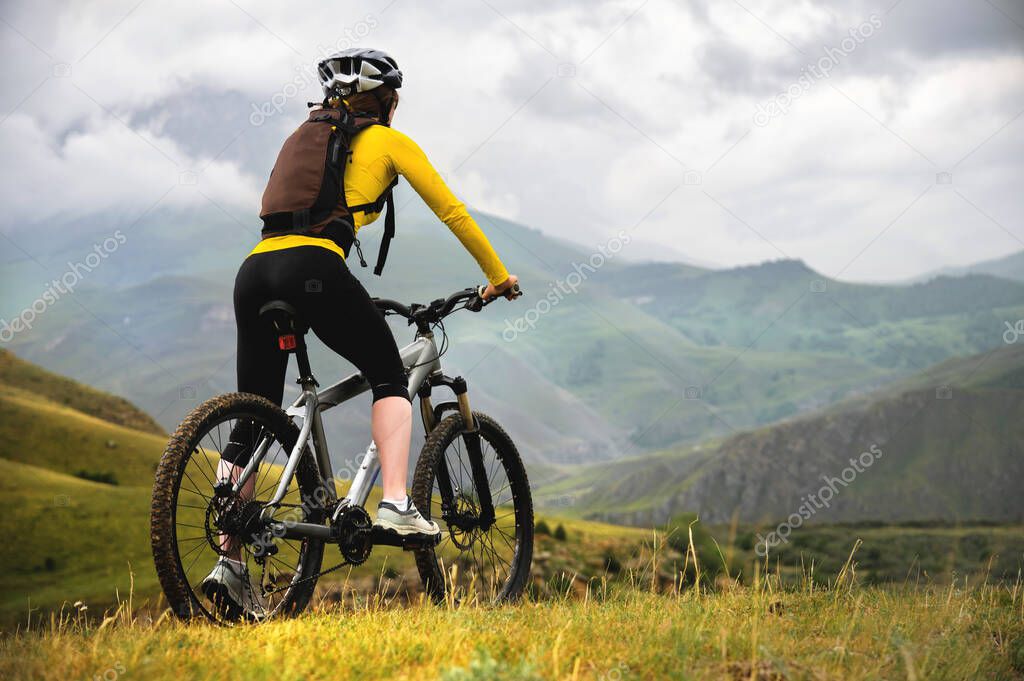 A young slender girl with a backpack and a bicycle helmet sits on a bicycle high in the mountains on a cloudy day and looks at the mountains and clouds.