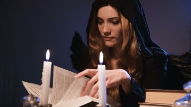 Medium close-up aantrekkelijk blank meisje met gouden lang haar in een zwarte kap zit aan een tafel en bladeren door een boek. Kostuum met vleugels. Kaarsen branden. Halloween viering concept — Stockvideo