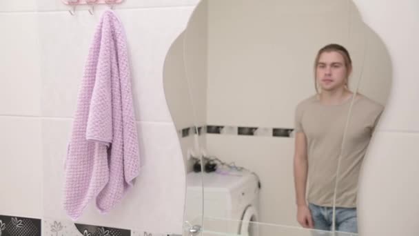 Un joven de pelo largo se acerca al espejo del baño y se mira a sí mismo por la mañana. El concepto de tratamientos matutinos en el baño — Vídeos de Stock