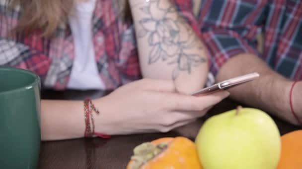 Młody uśmiech para facet i dziewczyna z długimi włosami siedzieć przy stole w kuchni i korzystać z telefonów komunikujących się z kimś w czatach i portalach społecznościowych. — Wideo stockowe