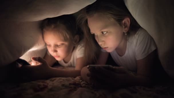 スマートフォンを持つ2人の女の子は毛布の下で寝ていません。子供の不眠症やデバイスやガジェットへのインターネット中毒 — ストック動画