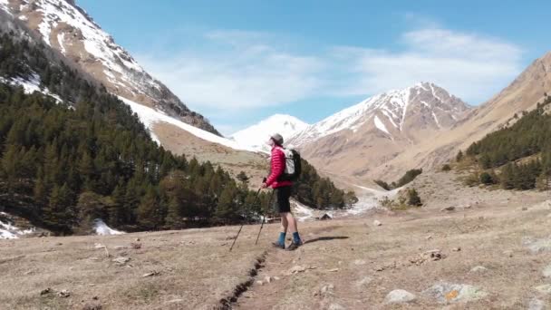 ショートサングラスの若い男とバックパック付きのキャップは、追跡棒で山の中のルートに沿って行きます。晴れた日には山の中でトレッキングやハイキング — ストック動画