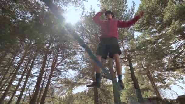 A young guy in a shorts jacket and a cap with sunglasses goes balancing on a stretched slackline in the coniferous forest in the afternoon. Outdoor sports and leisure concept — Stock Video