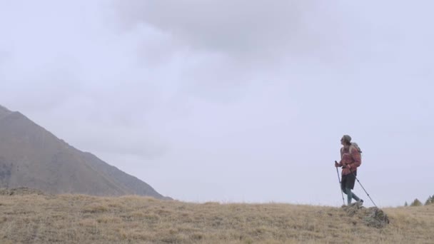 Un jeune homme grand en short chemise à carreaux dans une casquette et sac à dos en lunettes de soleil va sur un haut plateau sur fond de hautes montagnes par temps nuageux. Suivi et repos solitaire dans les montagnes — Video